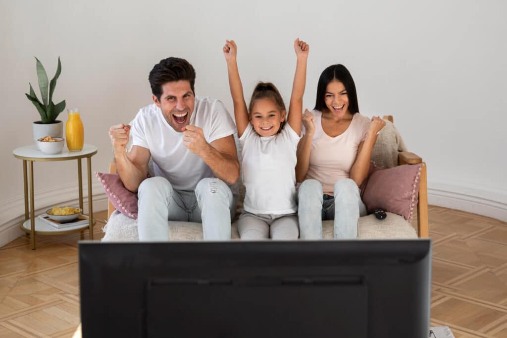 kids watching TV - A joyful family sitting together on a comfortable couch, with a smiling mom, dad, and their daughter watching television together, sharing a moment of happiness and connection.